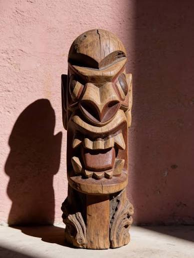 An unpainted carranca standing against a pink stucco wall in raking sunlight, throwing a hard shadow. The carving is angular: a domed head, deep-set slit eyes, a ridged snout, and a square mouth of blocky teeth.