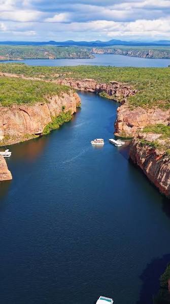 The São Francisco from above, winding deep blue between orange sandstone cliffs topped with green scrub. Two small tour boats sit near the bend, and blue hills fade out under a clouded sky.