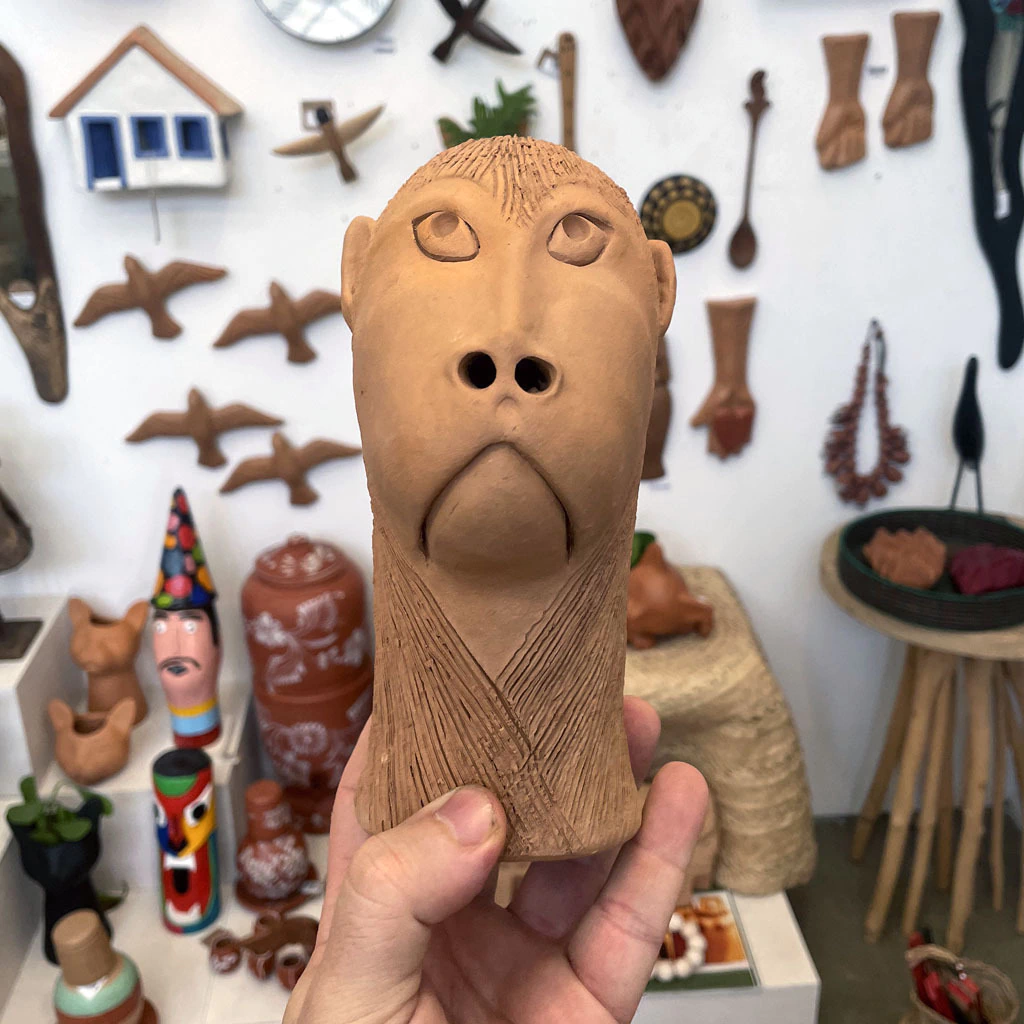A hand holding a small unglazed terracotta carranca: a long face with heavy-lidded eyes, two deep round nostrils and a downturned mouth, hair and chest scored with fine combed lines. A shop wall of clay birds and folk ceramics behind.