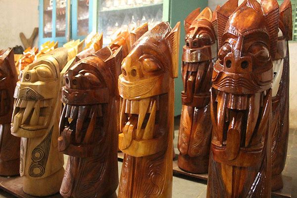 A row of unpainted carrancas in a shop, wood running from pale cream to dark red-brown. Each has a long snout and protruding fangs; the palest is cut all over with rows of circles and lines.
