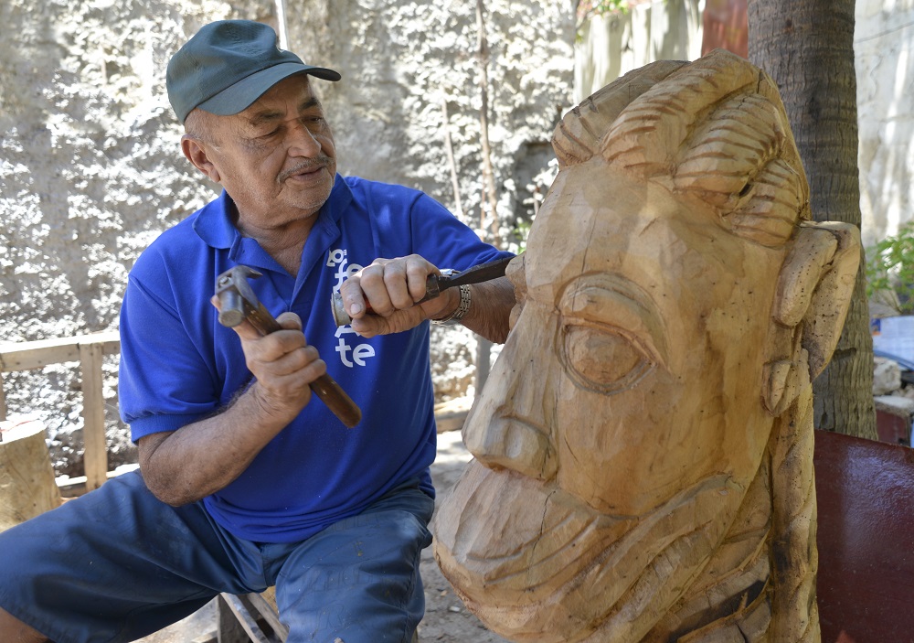 A carranqueiro in a blue polo shirt and cap cutting into a large half-finished carranca head with a mallet and chisel, outdoors in dappled light. The face is roughed out — brow, eye and ear already readable in the pale wood.